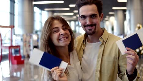 Excited-caucasian-couple-going-on-a-vacation-and-holding-passport-boarding-pass-at-airport.-Waving-with-check-in-passports,-smilling-to-the-camera,-embracing