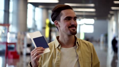 Happy,-handsome-bearded-american-man-showing-passport-with-flight-tickets-and-waving-it.-Looking-and-smilling-to-the-camera-while-standing-at-the-airport