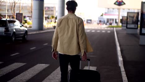 Confident,-tall-man-in-casual-with-the-black-baggage-walking-to-the-outside-airport-terminal-by-road.-Slow-motion.-Rare-view.-Blurred-backround