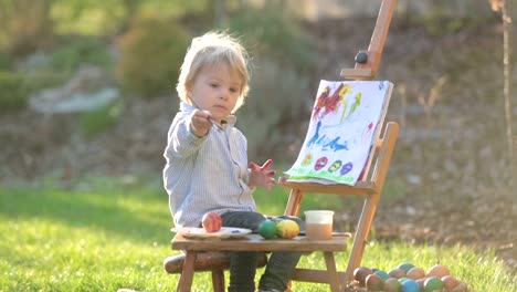 Niño-dulce,-pintando-huevos-en-el-jardín-con-polluelos-corriendo-a-su-alrededor-al-atardecer