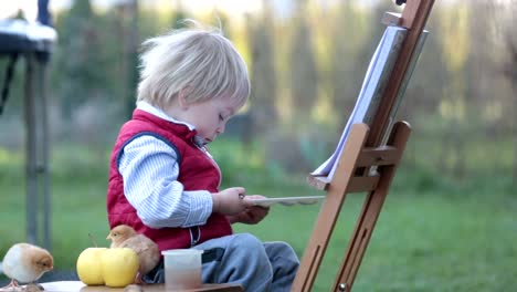 Niño-dulce,-pintando-huevos-en-el-jardín-con-polluelos-corriendo-a-su-alrededor-al-atardecer