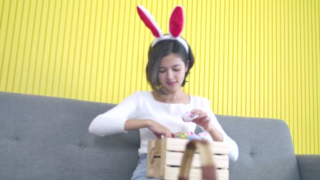 Easter,-holidays-and-people-concept.-Portrait-of-cheerful-happy-girl-wearing-bunny-ears-headband-sitting-on-the-sofa,-choosing-easter-eggs-decorated-colourful--of-handmade-in-wooden-crate-for-festival