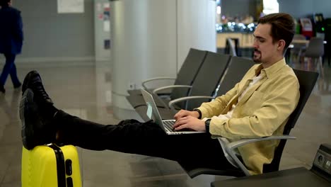 A-young-bored-man-is-sitting-at-the-airport,-waiting-for-the-plane-to-depart-and-using-a-laptop.-Typing-on-laptop,-holding-legs-on-his-yellow-suitcase.-Side-view