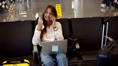 Elegante,-chica-bonita-con-camisa-blanca-utiliza-el-teléfono-y-la-computadora-portátil-en-el-aeropuerto-mientras-espera-el-embarque-en-la-sala-de-salida.-Hablando-con-amigos,-saqueando-y-emocionado.-Tener-el-pasaporte-en-la-mano