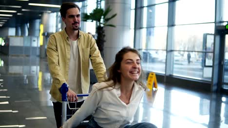 Romantic-couple-in-airport.-Attractive-young-woman-and-handsome-man-with-suitcases-are-ready-for-traveling.-Having-fun-on-luggage-trolley-while-waiting-for-departure.-Woman-immitating-flying,-smilling.-Slow-motion