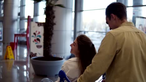 Funny-couple-in-airport.-Attractive-young-woman-and-handsome-man-with-suitcases-are-ready-for-traveling.-Having-fun-on-luggage-trolley-while-waiting-for-departure.-Woman-immitating-flying,-smilling,-man-running-and-pushing-trolley.-Rare-view