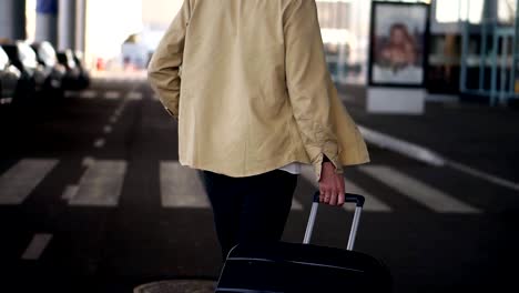 Close-up,-cropped-shot-of-married-man-with-the-black-baggage-walking-to-the-outside-airport-terminal-by-road.-Slow-motion.-Rare-view