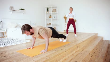 Young-couple-is-doing-push-ups-and-squats-excercises-at-home-in-cozy-bright-bedroom.-Stay-at-home-concept