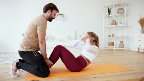 Young-couple-is-doing-sport-excercises-at-home-in-cozy-bright-bedroom.-Young-man-helps-his-girlfriend-to-do-abs-training.-Stay-at-home-concept