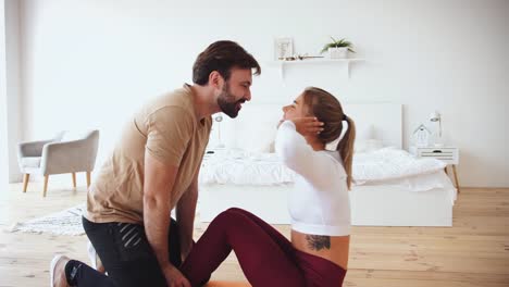 Young-couple-is-doing-sport-excercises-at-home-in-cozy-bright-bedroom.-Young-man-helps-his-girlfriend-to-do-abs-training.-Stay-at-home-concept