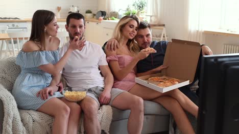 Group-of-friends-watching-TV-together-and-eating-pizza-at-home-on-sofa