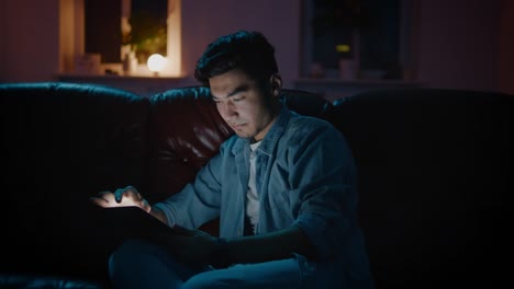 Glücklicher-und-positiver-asiatischer-Kerl-surfen-im-Internet-mit-Tablet-zu-Hause-auf-dem-Sofa.