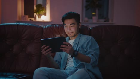 Glücklicher-asiatischer-Kerl,-der-Film-online-auf-Tablet-in-der-Nacht-sitzen-auf-dem-Sofa.