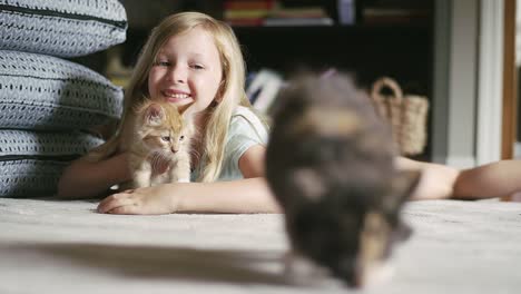 A-little-girl-laying-on-the-floor-holding-a-kitten-and-smiling,-with-another-kitten-in-the-foreground