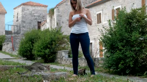 Young-woman-feeding-cat-standing-outdoor-in-summer-day.-She-carefully-pours-dry-food-out-of-glass