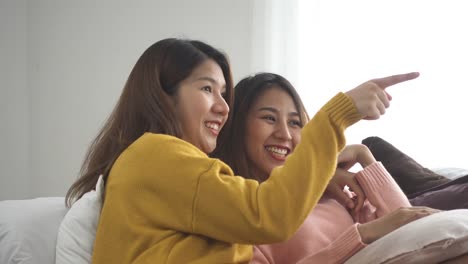 Feliz-pareja-lgbt-lesbiana-asiática-disfruta-del-entretenimiento-en-sala-de-estar.-Mujeres-hermosas-acostada-sobre-un-sofá-con-control-remoto-y-ver-video-y-películas-en-la-televisión-en-casa.