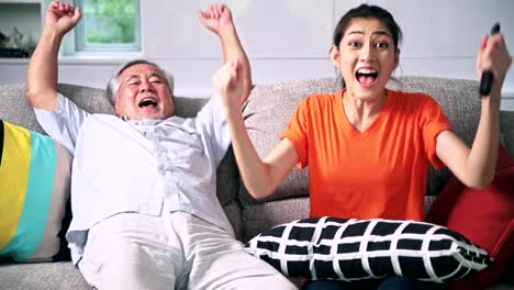 Old-man-watching-TV-with-his-daughter-in-living-room-in-early-evening.-Enjoying-entertainment-together,-senior-asian-man-and-young-woman.-Senior-lifestyle-family-concept.-Exciting-look.