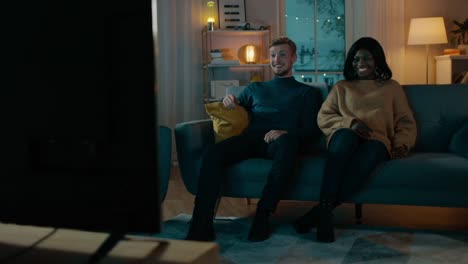 Happy-Diverse-Young-Couple-Watching-Comedy-on-TV-while-Sitting-on-a-Couch,-they-Laugh-and-Enjoy-Show.-Handsome-Caucasian-Boy-and-Black-Girl-in-Love-Spending-Time-Together-in-the-Cozy-Apartment.