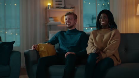 Happy-Diverse-Young-Couple-Watching-Comedy-on-TV-while-Sitting-on-a-Couch,-they-Laugh-and-Enjoy-Show.-Handsome-Caucasian-Boy-and-Black-Girl-in-Love-Spending-Time-Together-in-the-Cozy-Apartment.