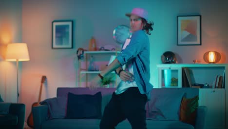 Handsome-Excited-Hip-Young-Man-is-Dancing-in-the-Living-Room-while-TV-Plays-in-the-Background.-He-is-Energetically-Moving-while-Screen-Adds-Reflections-on-Him.-Cozy-Room-is-Lit-with-Warm-Neon-Light.