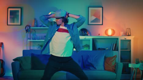 Handsome-Excited-Hip-Young-Man-is-Dancing-in-the-Living-Room-while-TV-Plays-in-the-Background.-He-is-Energetically-Moving-while-Screen-Adds-Reflections-on-Him.-Cozy-Room-is-Lit-with-Warm-Neon-Light.