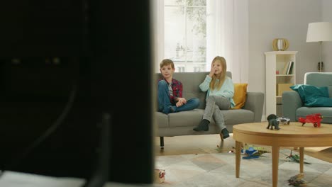 Zwei-niedliche-kleine-Mädchen-und-entzückende-junge-vor-dem-Fernseher-sitzen-auf-einem-Sofa.-Drei-glückliche-Kinder-Zeichentrickfilme-im-Fernsehen-und-Lachen-zu-Hause-im-Wohnzimmer.