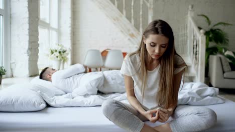 Depressed-young-woman-sitting-in-bed-and-crying-while-her-boylfriend-lying-in-bed-at-home