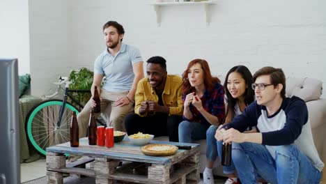 Grupo-étnico-multi-amigos-deportes-fans-viendo-el-evento-deportivo-en-la-televisión-juntos-comiendo-bocadillos-y-bebiendo-cerveza-en-su-casa-de-vacaciones