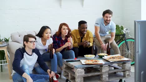 Gruppe-junger-Freunde-Sportübertragungen-im-Fernsehen-zusammen-Snacks-zu-essen-und-trinken-Bier-zu-Hause-passen.-Einige-von-ihnen-zufrieden-mit-ihrer-Mannschaft-gewinnen-aber-andere-enttäuscht