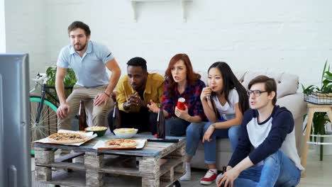 Gruppe-junger-Freunde-gerade-Olympischen-Spiele-Spiel-am-TV-zusammen-Snacks-zu-essen-und-Bier-zu-trinken.-Einige-von-ihnen-zufrieden-mit-ihrer-Mannschaft-gewinnen-aber-andere-enttäuscht