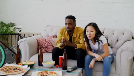 Multi-ethnic-couple-watching-sports-game-on-TV-together-at-home-and-yelling-while-their-favourite-team-win-at-home