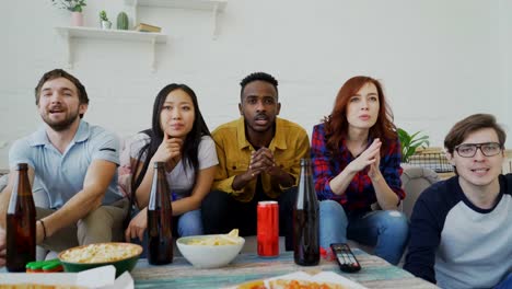 Grupo-de-jóvenes-amigos-viendo-los-Juegos-Olímpicos-coinciden-en-TV-juntos-comiendo-bocadillos-y-bebiendo-cerveza.-Algunos-de-ellos-felices-con-su-equipo-ganando-pero-hombre-decepcionado