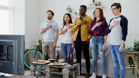 Grupo-étnico-multi-de-amigos-deporte-aficionados-cantando-el-himno-nacional-antes-de-ver-el-Campeonato-de-deporte-en-la-televisión-juntos-en-casa