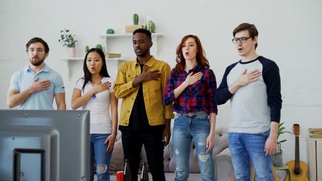 Multi-ethnic-group-of-friends-sport-fans-singing-national-anthem-before-watching-sports-championship-on-TV-together-at-home