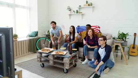 Grupo-étnico-multi-de-amigos-con-banderas-nacionales-de-Estados-Unidos-viendo-el-Campeonato-del-deporte-en-la-televisión-animando-a-su-equipo-favorito-en-casa