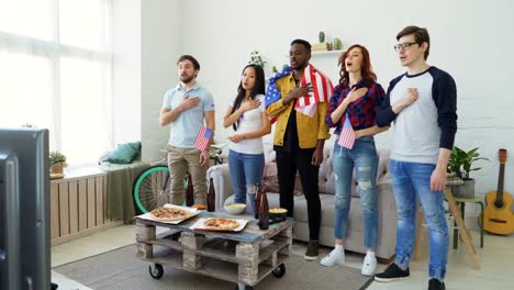 Multi-étnico-grupo-de-amigos-aficionados-cantando-el-himno-nacional-de-Estados-Unidos-antes-de-ver-el-Campeonato-de-deportes-en-la-televisión-juntos-en-la-casa-del-deporte