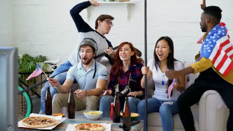 Multi-ethnic-group-of-friends-sports-fans-with-USA-national-flags-watching-winter-olympic-games-on-TV-together-cheering-up-favourite-team-at-home