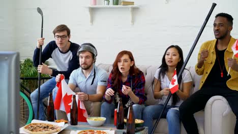 Grupo-de-jóvenes-amigos-deportes-aficionados-con-banderas-nacionales-canadienses-ver-Campeonato-del-deporte-en-la-televisión-juntos-animando-hasta-equipo-favorito-en-casa