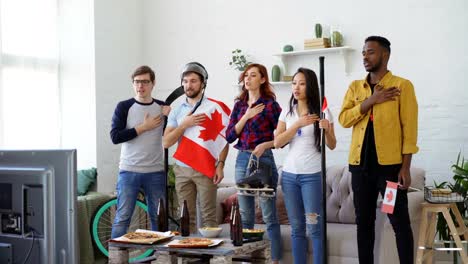 Multi-ethnischen-Gruppe-von-Freunden-hören-und-singen-kanadischen-Nationalhymne-vor-gerade-Sport-Meisterschaft-im-Fernsehen-zusammen-zu-Hause