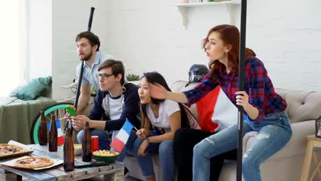 Multi-étnico-grupo-de-amigos-fans-de-los-deportes-con-banderas-nacionales-francesas,-viendo-el-Campeonato-de-hockey-en-la-televisión-animando-a-su-equipo-favorito-en-casa