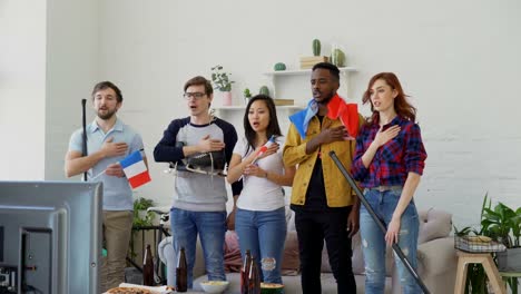 Multi-ethnic-group-of-friends-listening-and-singing-French-national-anthem-before-watching-sports-championship-on-TV-together-at-home