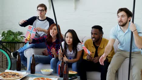 Group-of-young-friends-sports-fans-with-Norwegian-national-flags-watching-sport-championship-on-TV-together-and-happy-about-winning-favourite-team-at-home