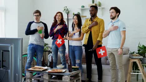 Multi-ethnic-group-of-friends-listening-and-singing-Swiss-national-anthem-before-watching-sports-championship-on-TV-together-at-home