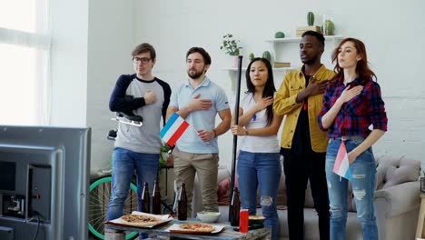 Multi-ethnic-group-of-friends-listening-and-singing-Dutch-national-anthem-before-watching-sports-championship-on-TV-together-at-home