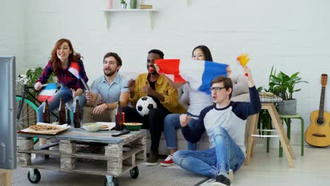 Multi-ethnic-group-of-friends-sports-fans-with-French-flags-watching-football-championship-on-TV-together-at-home-and-cheering-up-favourite-team