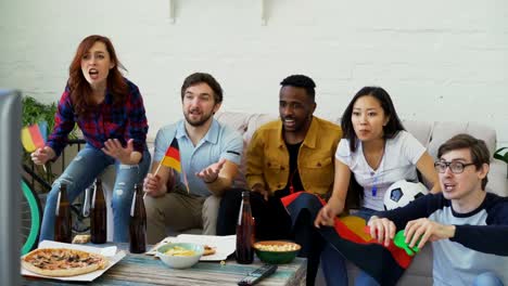 Multi-ethnic-group-of-friends-sports-fans-with-German-flags-watching-football-championship-on-TV-together-at-home-and-cheering-up-favourite-team