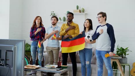 Multi-ethnischen-Gruppe-von-Freunden-hören-und-singen-der-deutschen-Nationalhymne-vor-gerade-Sport-Meisterschaft-im-Fernsehen-zusammen-zu-Hause