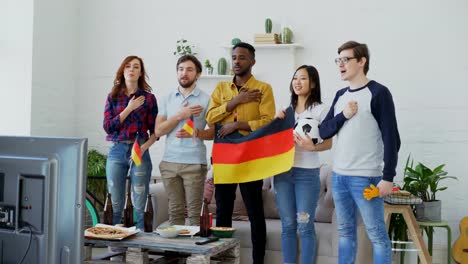 Multi-ethnic-group-of-friends-listening-and-singing-German-national-anthem-before-watching-sports-championship-on-TV-together-at-home