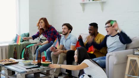 Multi-ethnic-group-of-friends-sports-fans-with-German-flags-watching-football-championship-on-TV-together-at-home-and-cheering-up-favourite-team