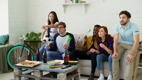 Multi-ethnic-group-of-friends-sports-fans-with-Brazilian-flags-watching-football-championship-on-TV-together-at-home-and-cheering-up-favourite-team